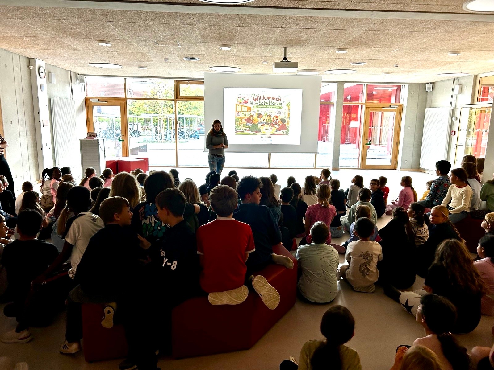 Viele Kinder sitzen in der Aula auf dem Boden und auf roten Sitzelementen und schauen zur Leinwand, auf der die Begrüßung zum Schulforum gezeigt wird.